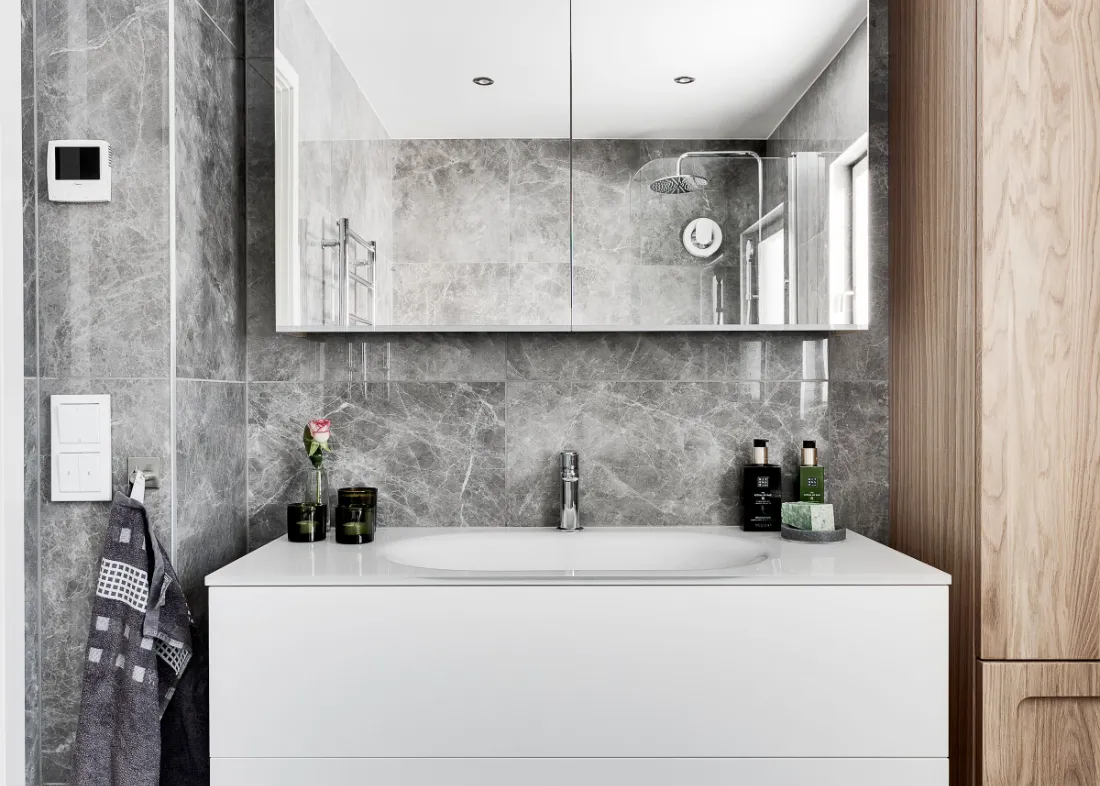 Mueble suspendido bajo lavabo contra una pared de piedra gris, con espejo y aplique encima.