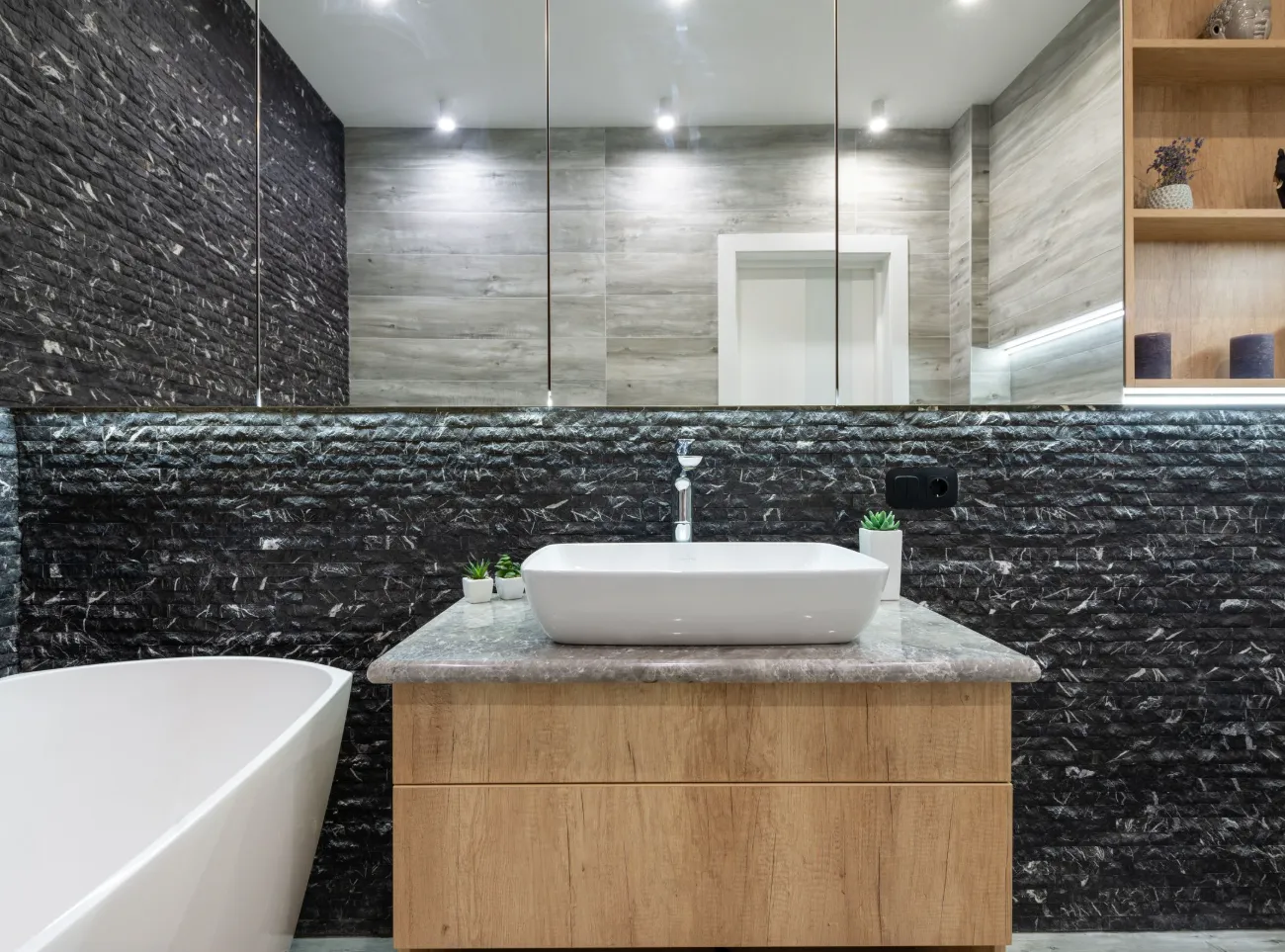 Baño oscuro con pared de pizarra, lavabo sobre encimera de madera y bañera exenta.