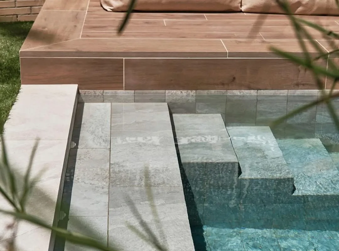Borde de una piscina terminada: banco de obra revestido en cerámica efecto madera con cojines, muro de ladrillo visto y el agua al fondo.