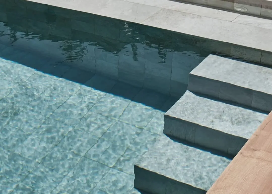 Piscina terminada vista desde arriba, con los peldaños y el vaso revestidos en piedra gris verdosa y una esquina de pavimento cerámico efecto madera.